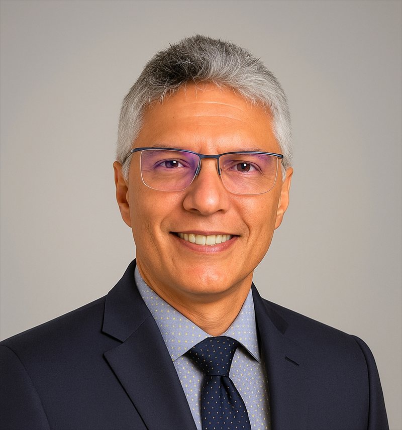 Retrato de José Gonçalves de Souza Júnior, de terno azul-marinho e óculos, sorrindo, em fundo cinza claro.